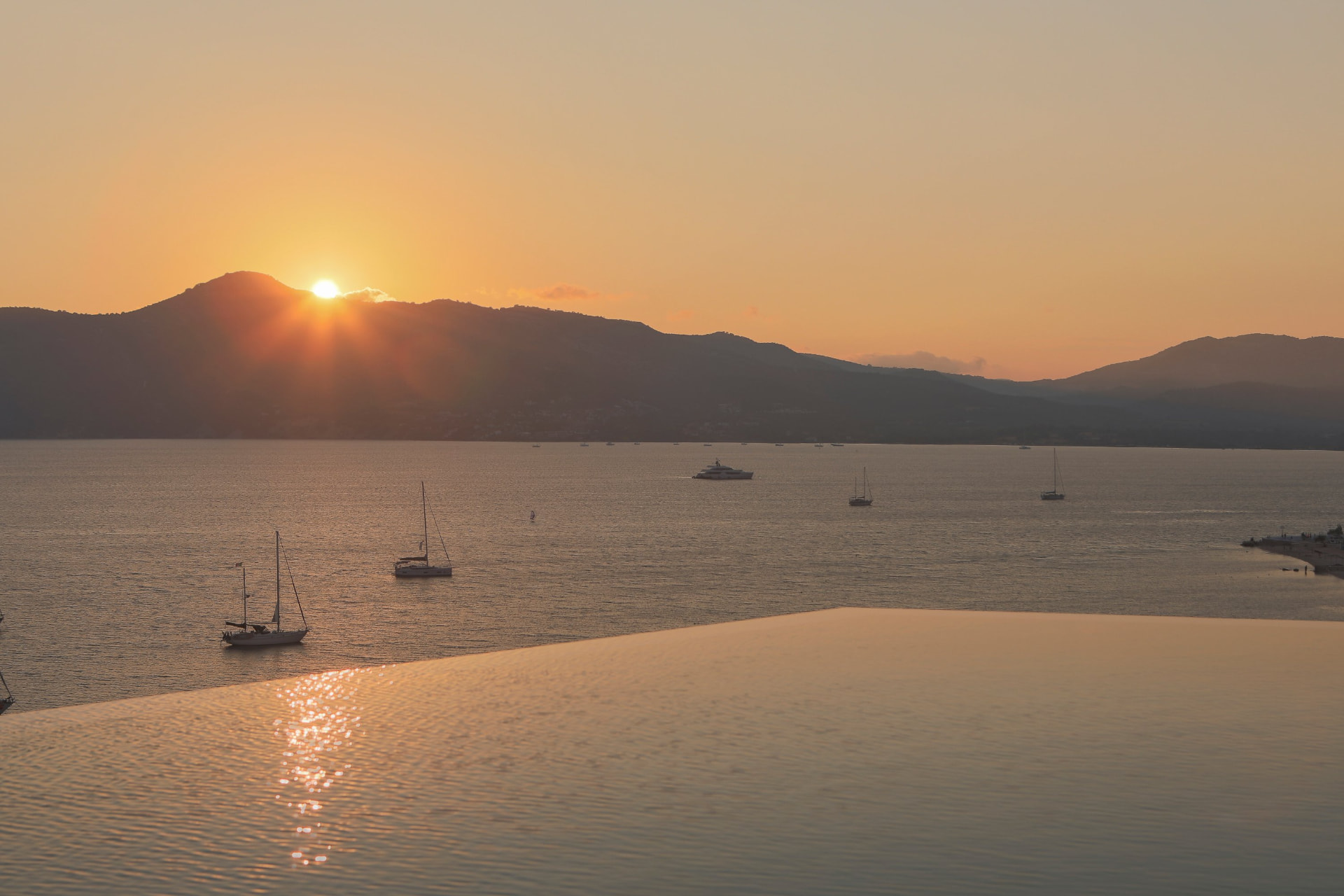 Sunset over the mountains reflected in the infinity pool with yachts in the bay