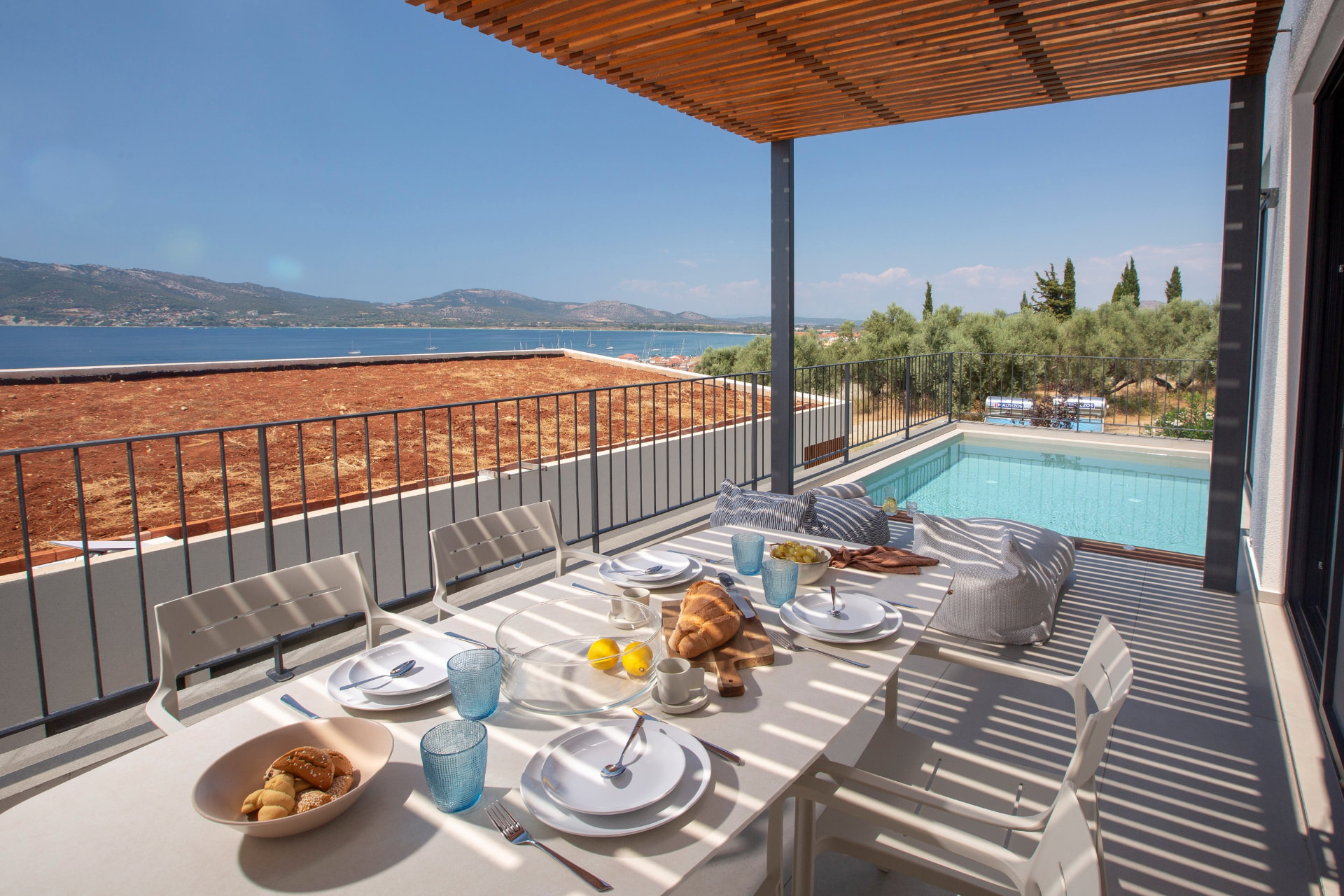 Pergola-shaded balcony dining table set for breakfast beside a private pool with bay views